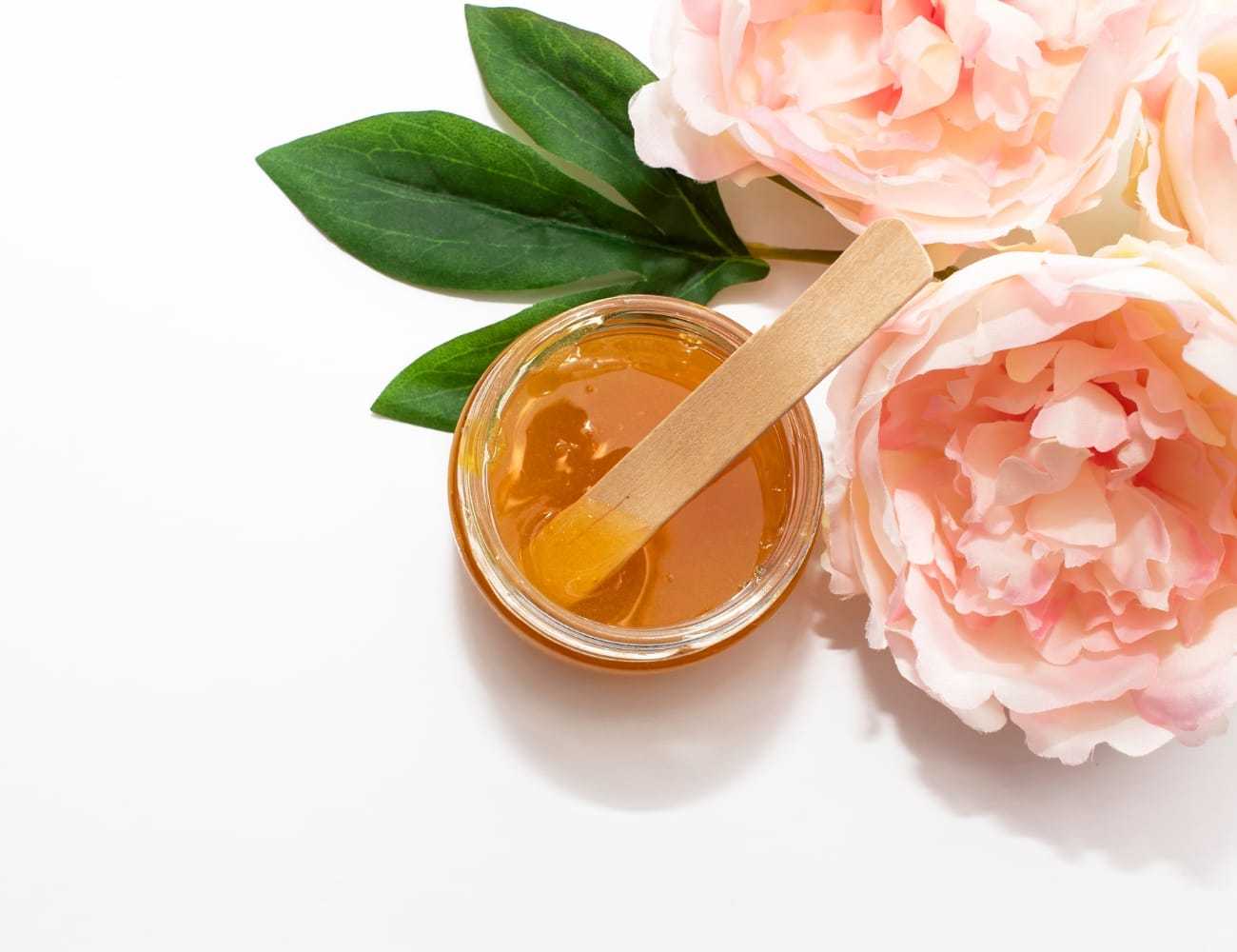 Jar of honey with wooden stick next to pink flowers and green leaves on a white background.