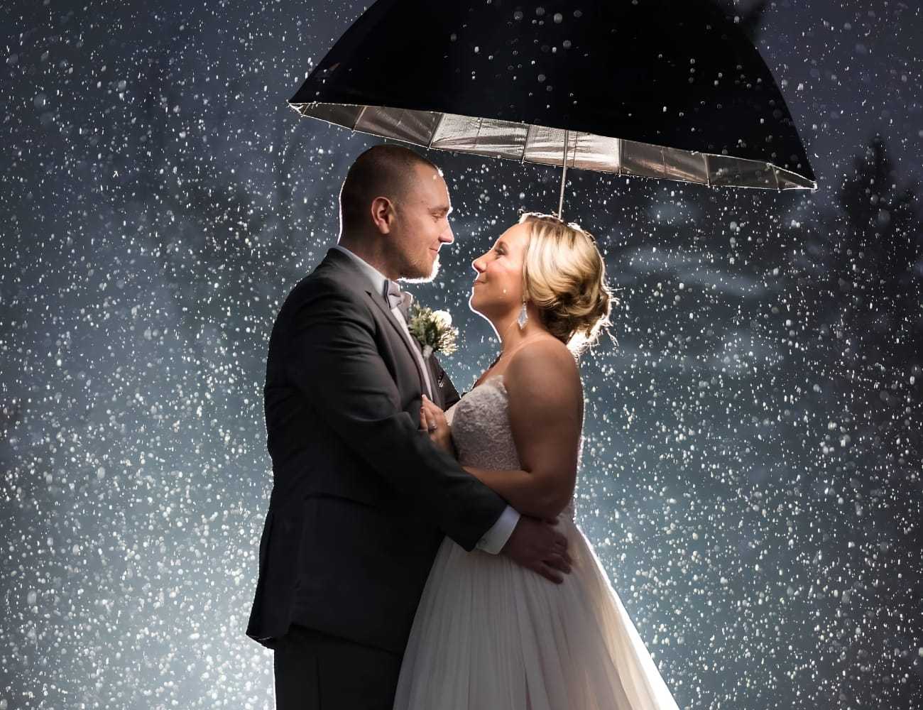 Couple embraces under umbrella in snowy evening, bride in white dress, groom in gray suit.